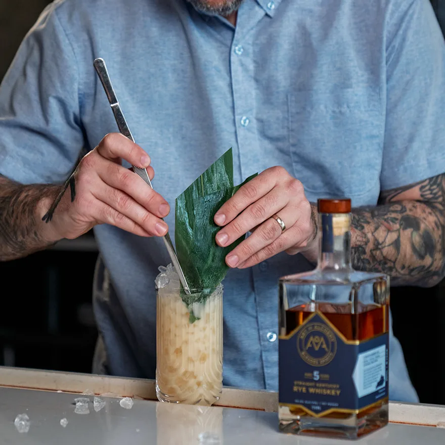 Bird Rights being finished in a Tom Collins glass packed with pebble ice, a broad green leaf set into the top, beside a bottle of Art of Alchemy 5-Year Straight Kentucky Rye.