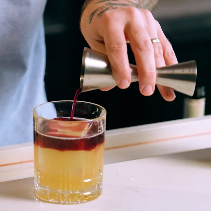 A New York Sour in a rocks glass, red Malbec being poured from a jigger to float in a band across the pale citrus base.
