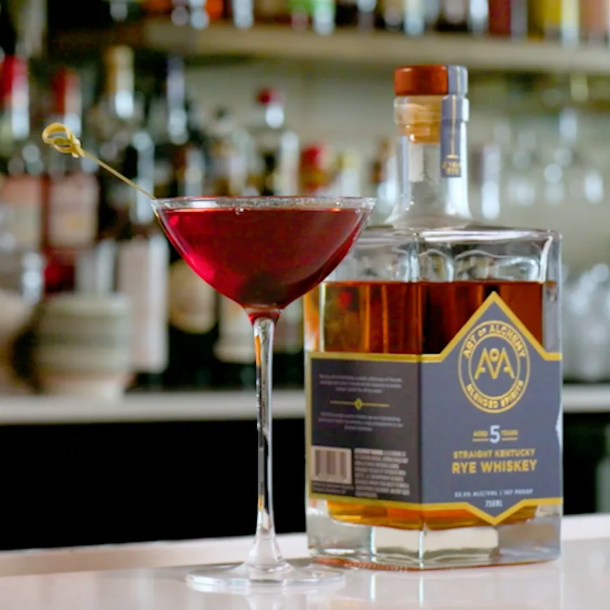 The Backdrop served up in a coupe, deep red and garnished with a cocktail cherry, beside a bottle of Art of Alchemy 5-Year Straight Kentucky Rye.