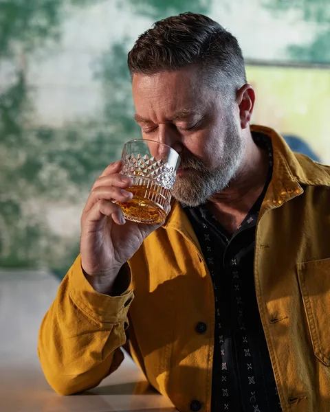 A guest at the bar draws in the aroma from a cut-crystal rocks glass of whiskey, a weathered green wall behind.
