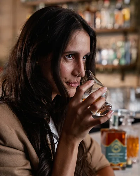 A guest turns from the bar mid-sip, a neat pour in a tulip glass, shelves of bottles out of focus behind.