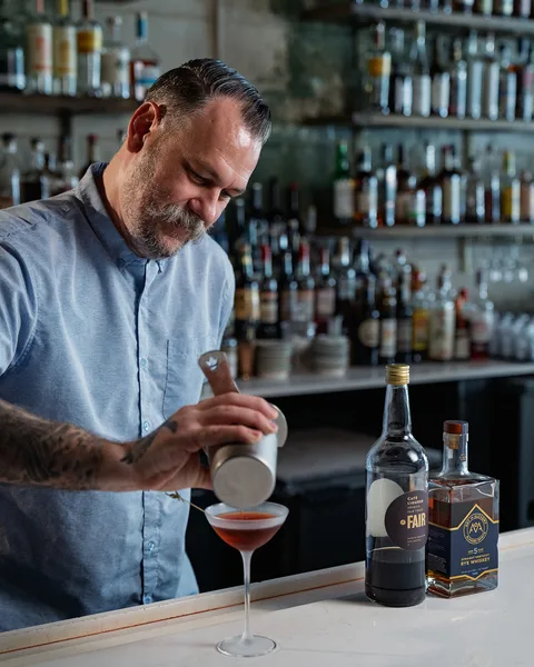 A bartender strains a cocktail into a stemmed glass, a bottle of Art of Alchemy rye and a bottle of café liqueur on the bar beside.